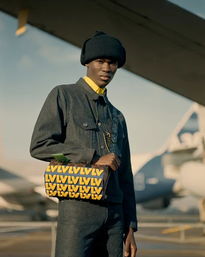 A stylish African American man in a denim jacket and black beanie poses with a Louis Vuitton bag in front of an airplane.