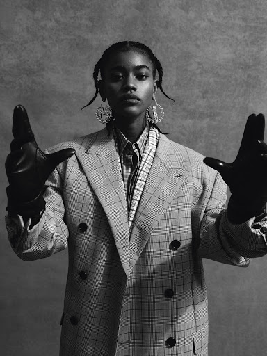 Black woman wearing a plaid blazer and leather gloves with hands raised against a textured backdrop
