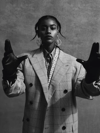 Black woman wearing a plaid blazer and leather gloves with hands raised against a textured backdrop