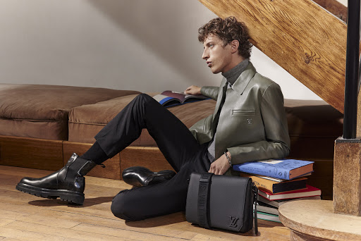A young adult male model in an olive jacket is seated on a floor with books and a luxury Louis Vuitton messenger bag.