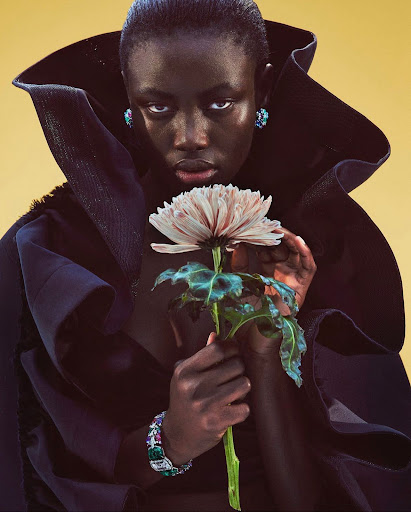 Striking portrait of a dark-skinned woman with statement collar and jewelry, holding a flower against a light yellow backdrop in a studio.
