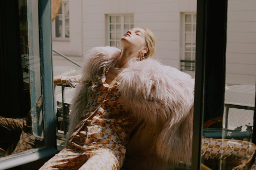 A blonde adult woman in a patterned jumpsuit and pink fur coat reclines in a window with a sunlit exterior. Editorial fashion photography.