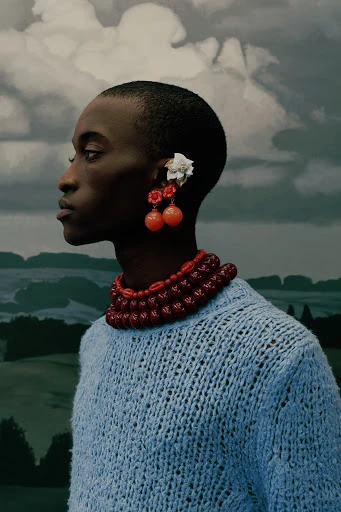Portrait of an African American person in a blue knit sweater with cherry earrings and a beaded necklace, set against a cloudy sky backdrop.