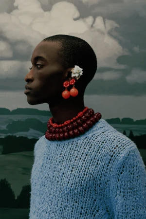 Portrait of an African American person in a blue knit sweater with cherry earrings and a beaded necklace, set against a cloudy sky backdrop.