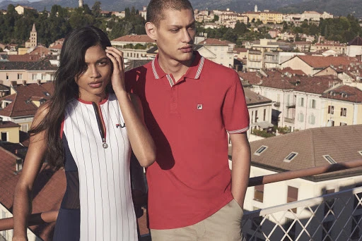 Two models, an adult woman and an adult male, pose on a rooftop in athletic wear, with an urban skyline and blue sky in the background.