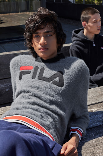 Two men wear FILA branded apparel outdoors. The first man wears a grey sweater with the FILA logo, while the second man wears a black FILA hoodie.