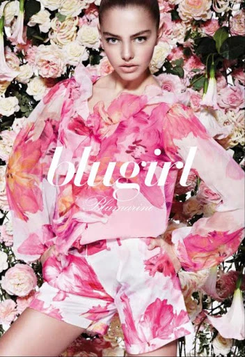 Fashion shot of a young woman in a floral print shirt and shorts in front of a backdrop of roses, promoting the brand Blugirl.