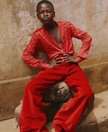 Young Black boy sitting on a rock wearing a red patterned shirt and red pants poses outdoors with hands on hips in natural light.