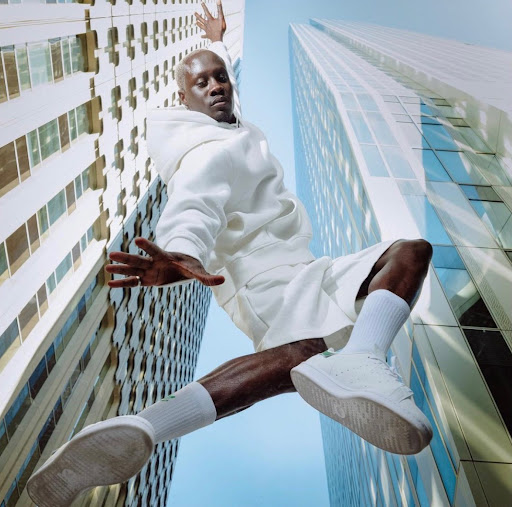 Bald man jumps in the air wearing a white hoodie, shorts, socks, and sneakers in an urban environment with tall buildings.