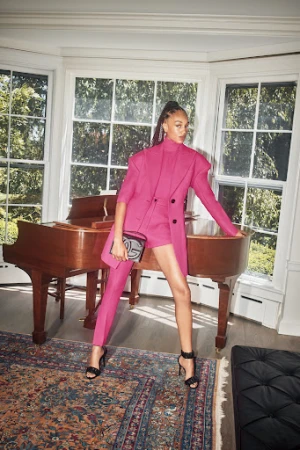 Woman poses in a pink blazer and shorts with high heels, holding a bag, inside a home with a piano and a patterned rug.