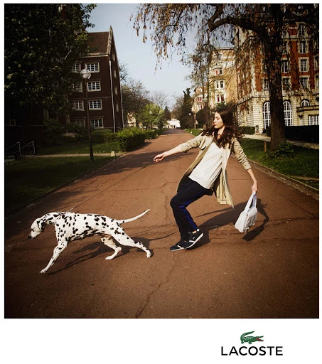 Fashion shot of a woman and Dalmatian dog walking down a tree-lined street with brick buildings and the Lacoste logo in the corner.