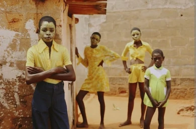 Four children wearing yellow and green outfits with decorative facial makeup standing outdoors near a brick wall.