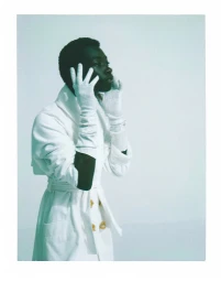 Black man wearing a white belted coat and white gloves with hands framing his face against a pale background