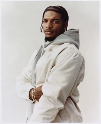 Portrait of a black man with braided hair wearing a light jacket over a grey hoodie against a plain light background