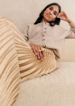 South Asian woman with dark hair seated on a sofa wearing a beige cardigan and pleated skirt with gold rings.