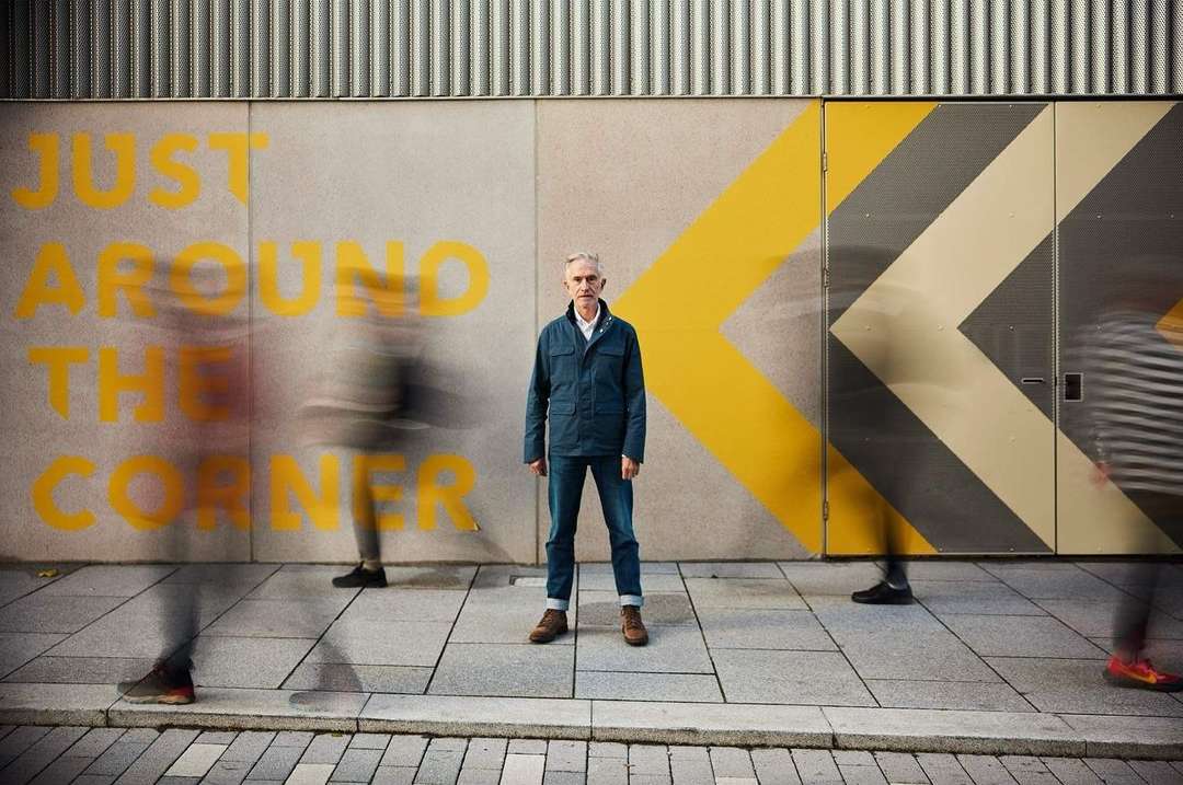 Adult man standing in front of wall with yellow directional arrows and 'Just Around The Corner' painted on it, blurred pedestrians pass by.