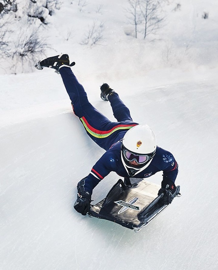Person in blue racing suit and helmet sledding down snowy hill, captured in an eye-level wide shot, outdoor scene with trees in the distance.