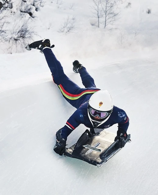 Person in blue racing suit and helmet sledding down snowy hill, captured in an eye-level wide shot, outdoor scene with trees in the distance.