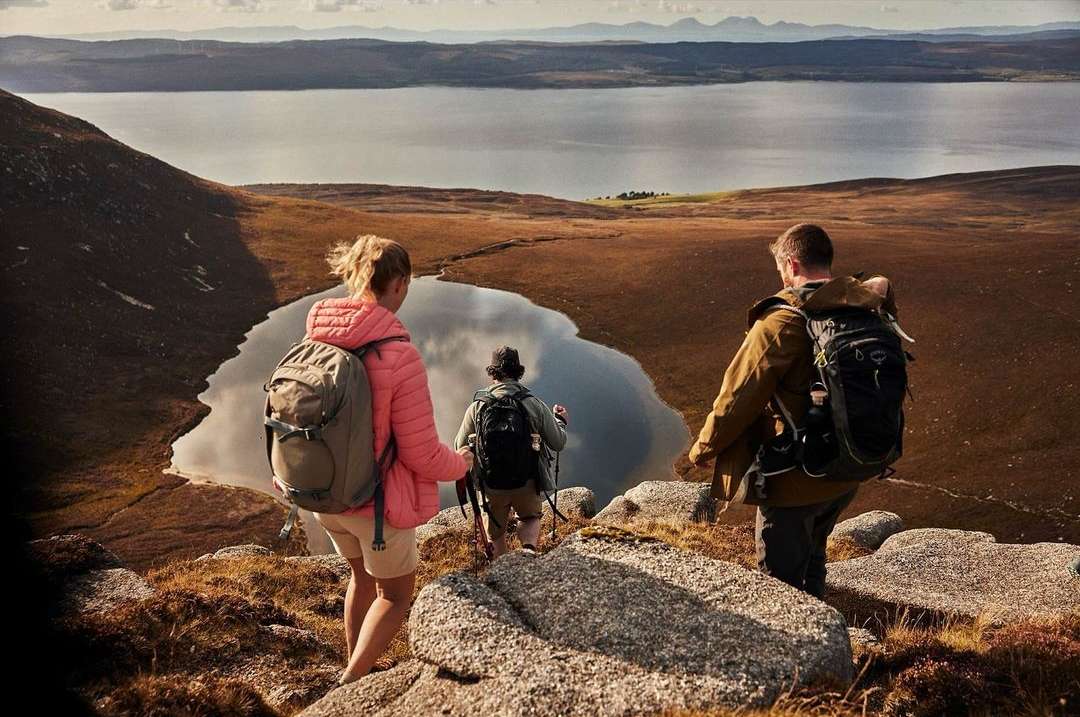 Three hikers, two with backpacks, walk downhill on a rocky path toward a valley and lake with mountains on the horizon.