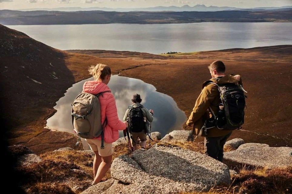 Three hikers, two with backpacks, walk downhill on a rocky path toward a valley and lake with mountains on the horizon.