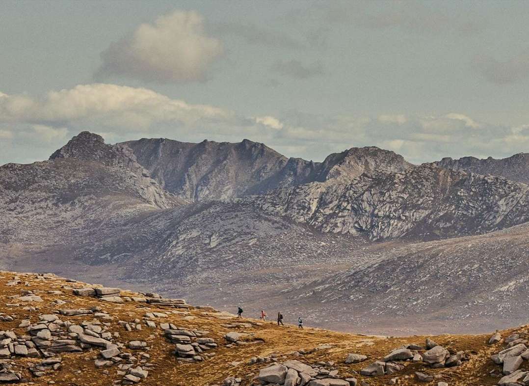 Scenic mountain range landscape with multiple hikers traversing a rocky terrain under a cloudy sky, creating a sense of adventure and scale.