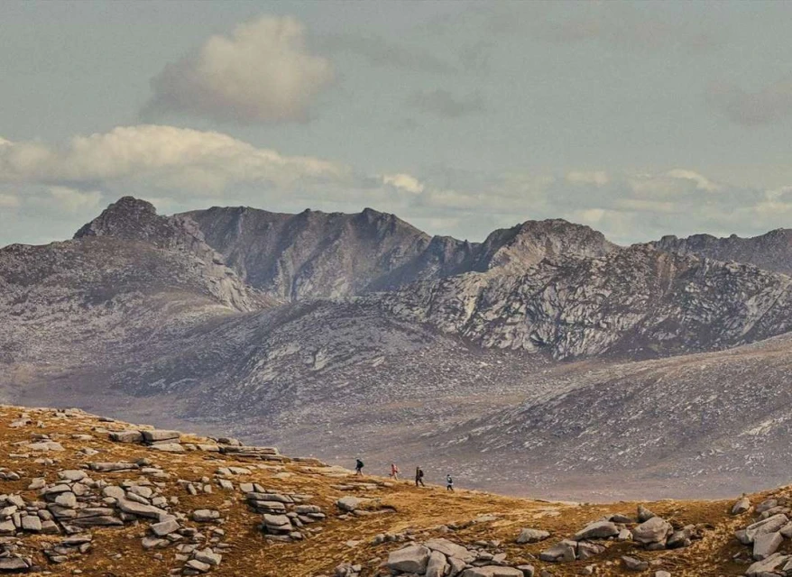 Scenic mountain range landscape with multiple hikers traversing a rocky terrain under a cloudy sky, creating a sense of adventure and scale.