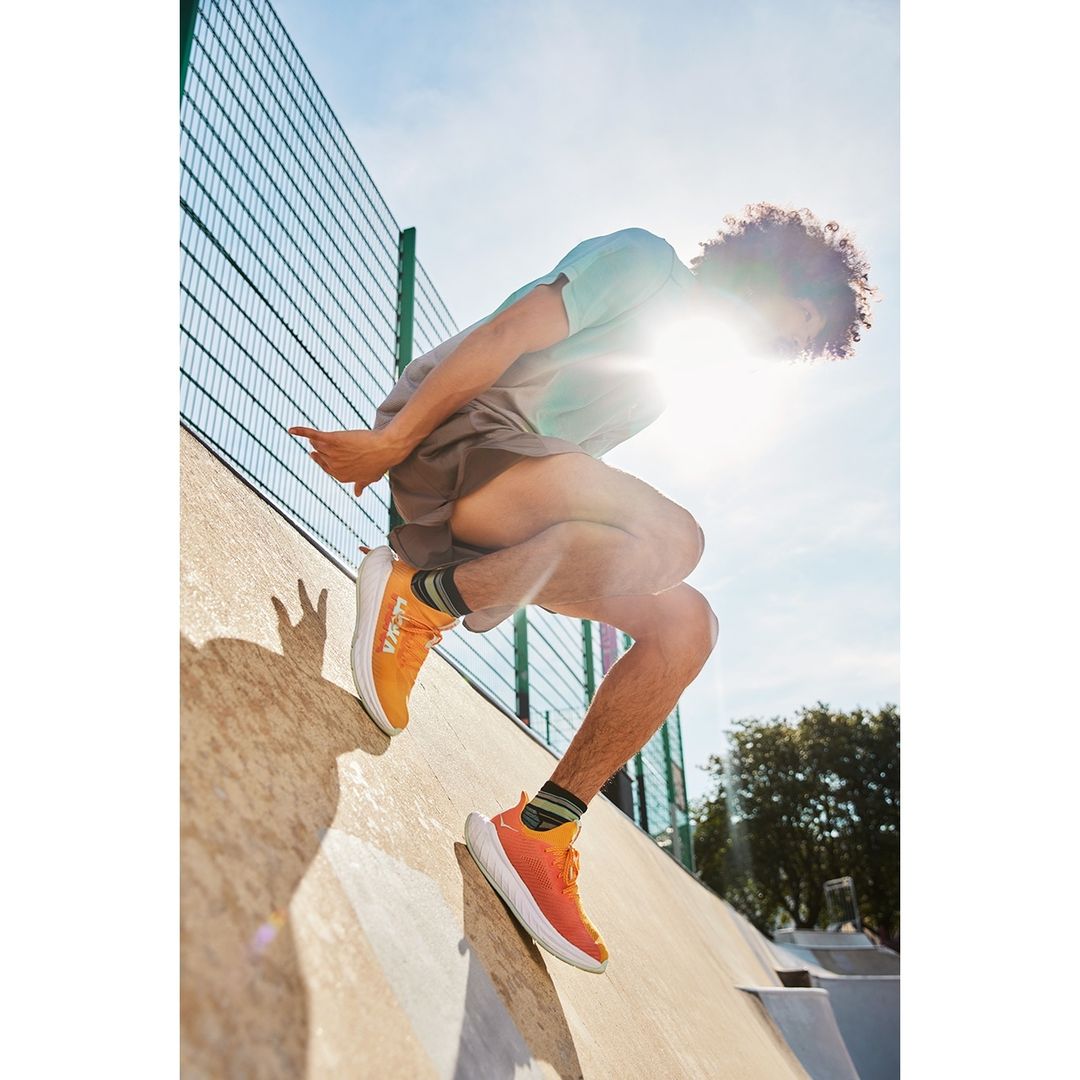 An athletic male with curly hair jumps on an outdoor concrete ramp wearing orange running shoes, a t-shirt, and shorts on a sunny day.
