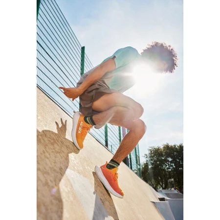 An athletic male with curly hair jumps on an outdoor concrete ramp wearing orange running shoes, a t-shirt, and shorts on a sunny day.