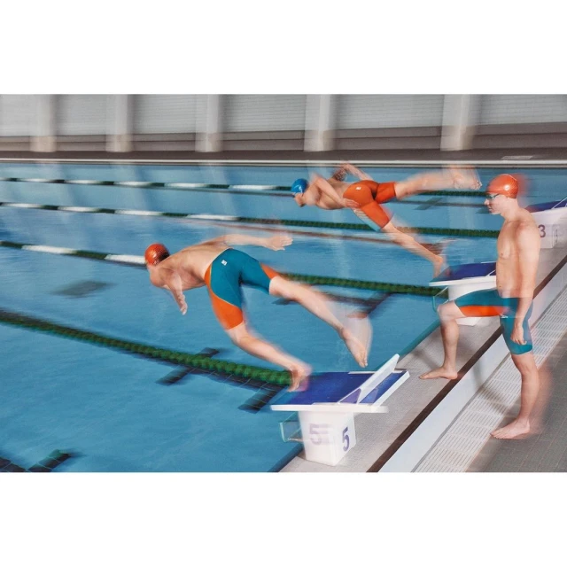 A group of male swimmers diving into a blue pool in an indoor swimming pool competition with motion blur and a bright setting.