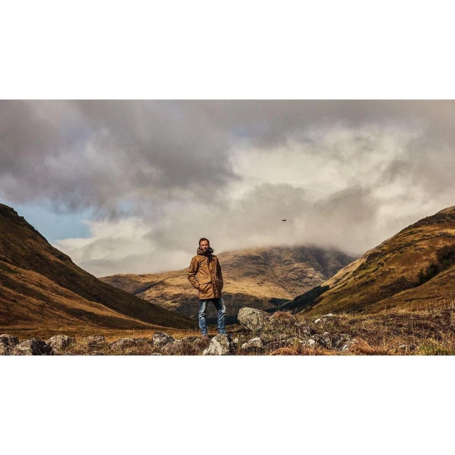Adult man stands among rocks in a valley with grassy hills under a cloudy sky, creating an outdoor scenic view.
