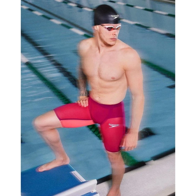 Male swimmer in cap and goggles standing poolside in red and maroon racing suit; Speedo logo visible; blurred motion effect; water background.