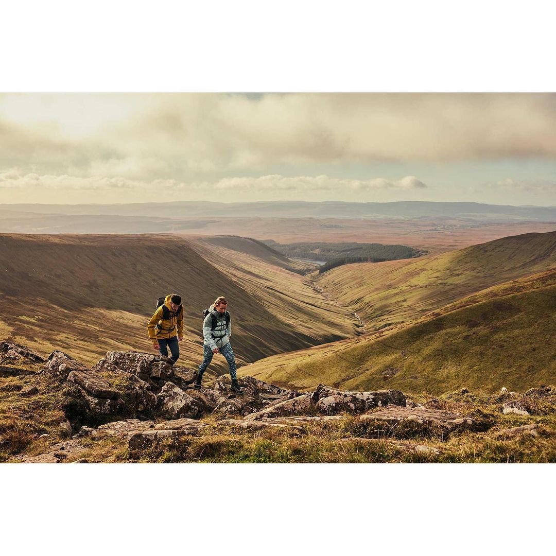 Two people hike along a rocky ridge in a mountain valley landscape on a cloudy day, enjoying nature and outdoor activity.