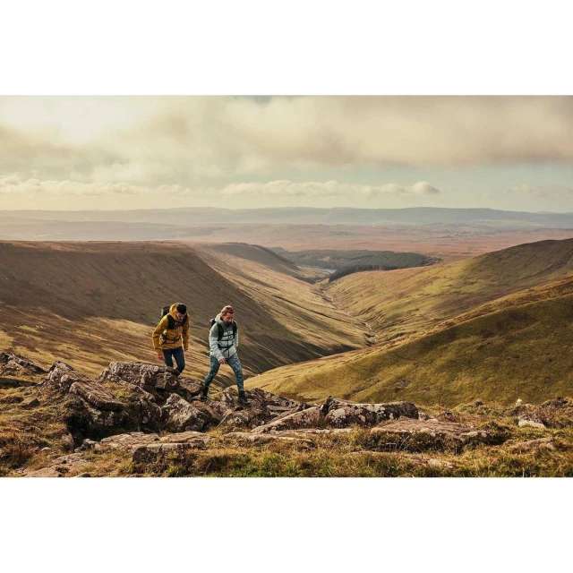 Two people hike along a rocky ridge in a mountain valley landscape on a cloudy day, enjoying nature and outdoor activity.