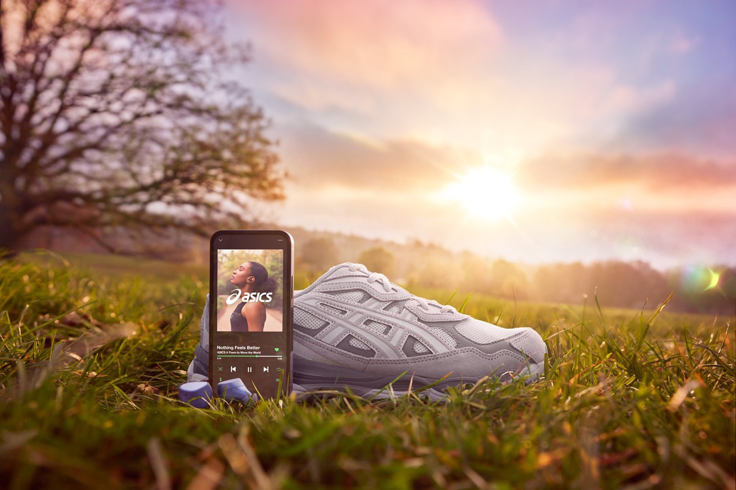 Running shoe and phone with streaming app on a grassy field at sunset. Asics logo displayed on the phone's screen. Outdoor setting.