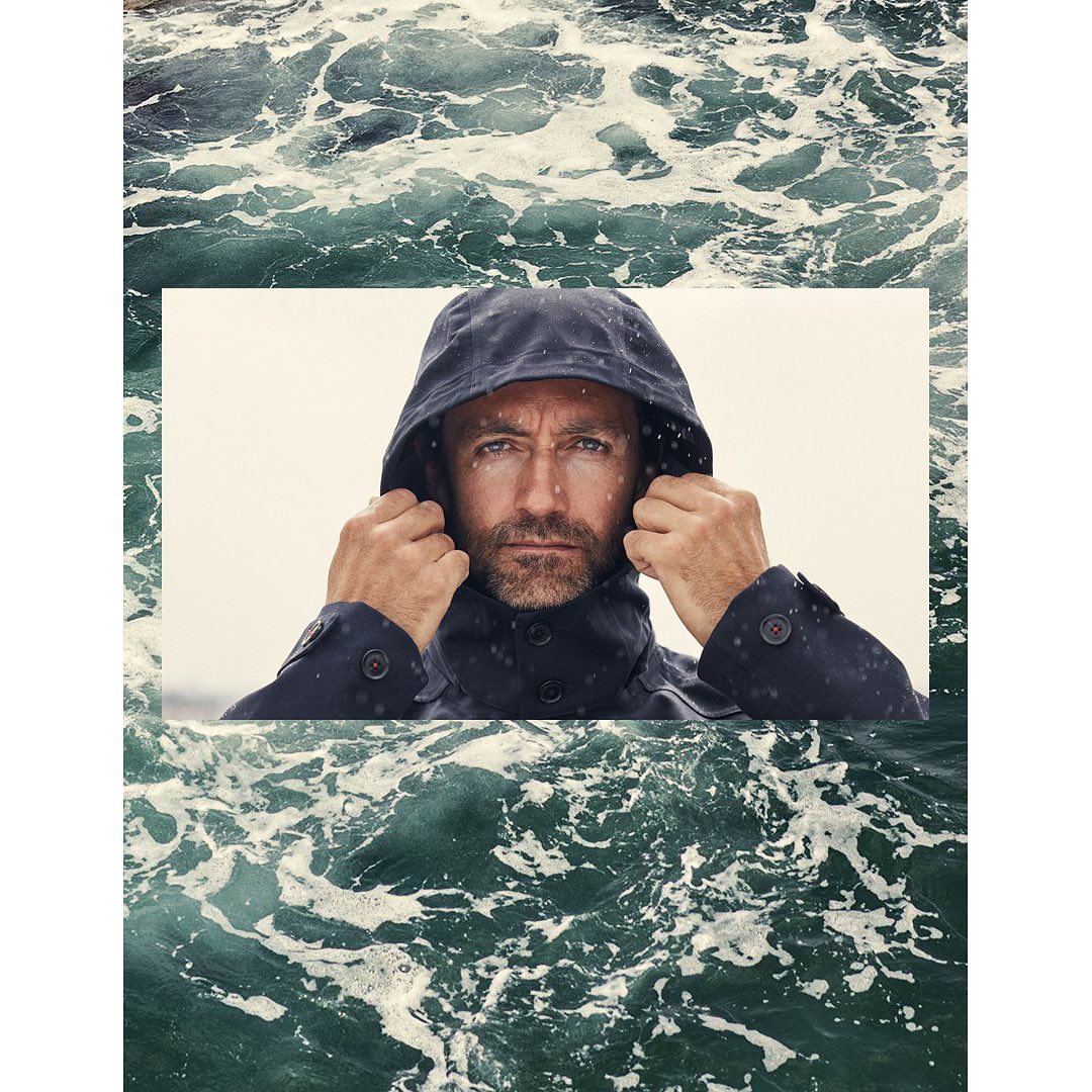 Man in hooded jacket gazes forward against a backdrop of swirling water. A portrait with fashion and nature elements.
