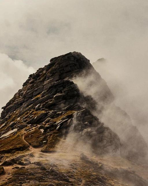 Rocky mountaintop shrouded in mist and low clouds, illuminated by soft, diffused light on a cloudy day.