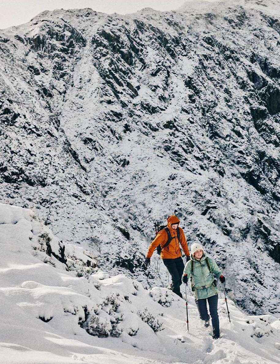 Two people hike a snow-covered mountain trail with trekking poles under overcast skies, surrounded by winter mountain scenery.