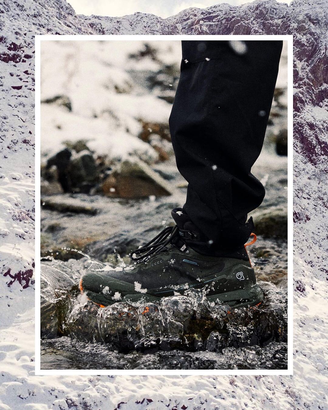 Hiking shoe steps on river rock in a mountain stream. Person in black hiking pants. Snowy mountain background. Outdoor environment.
