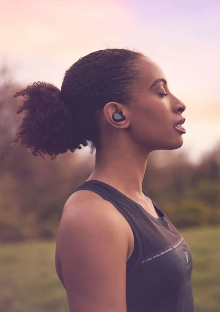 Side profile of a Black woman with curly hair wearing earbuds and a sleeveless top, outside in a blurry green and yellow field.