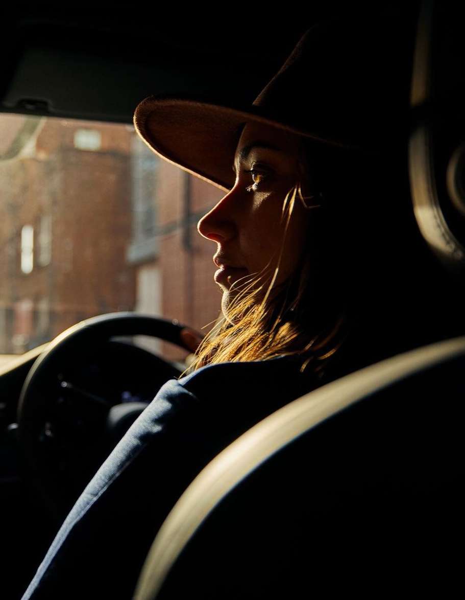 Adult woman wearing hat drives car during daytime. Sunlight illuminates her face in side-view portrait. Vehicle interior is dark.