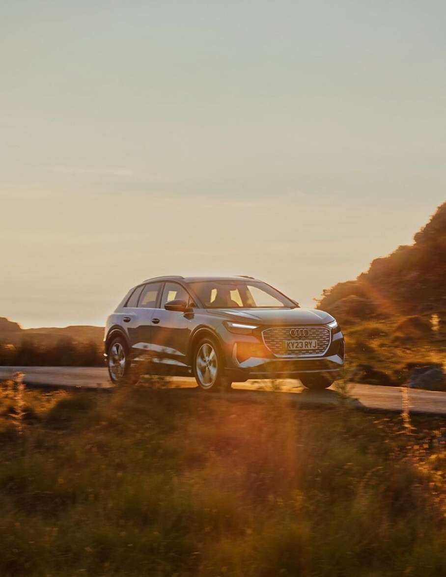 Gray Audi Q4 e-tron SUV driving on a road through a grassy hillside at sunset with a sun flare in the background.