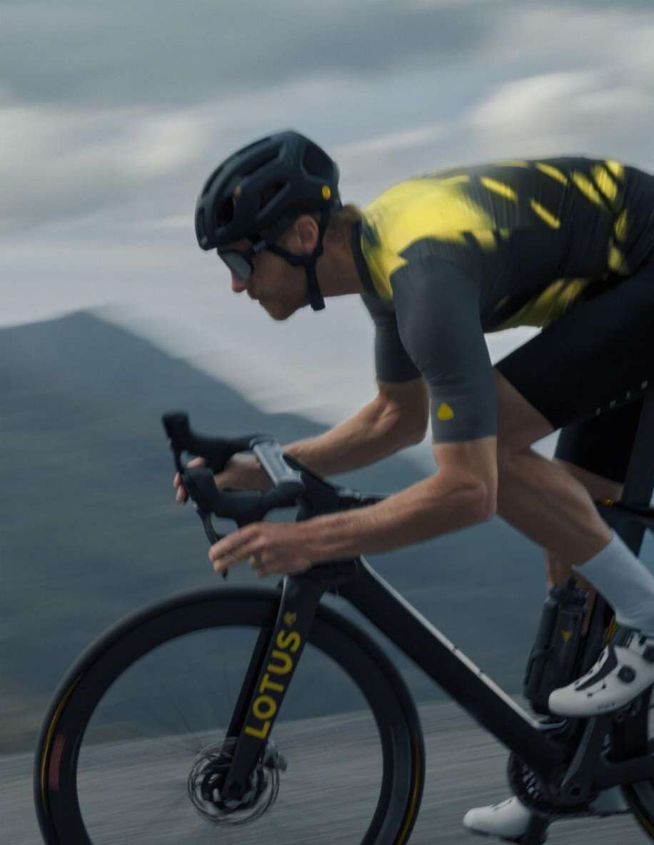 Adult male cyclist riding a black Lotus bicycle on a paved road with a gray mountain background wearing a black helmet.