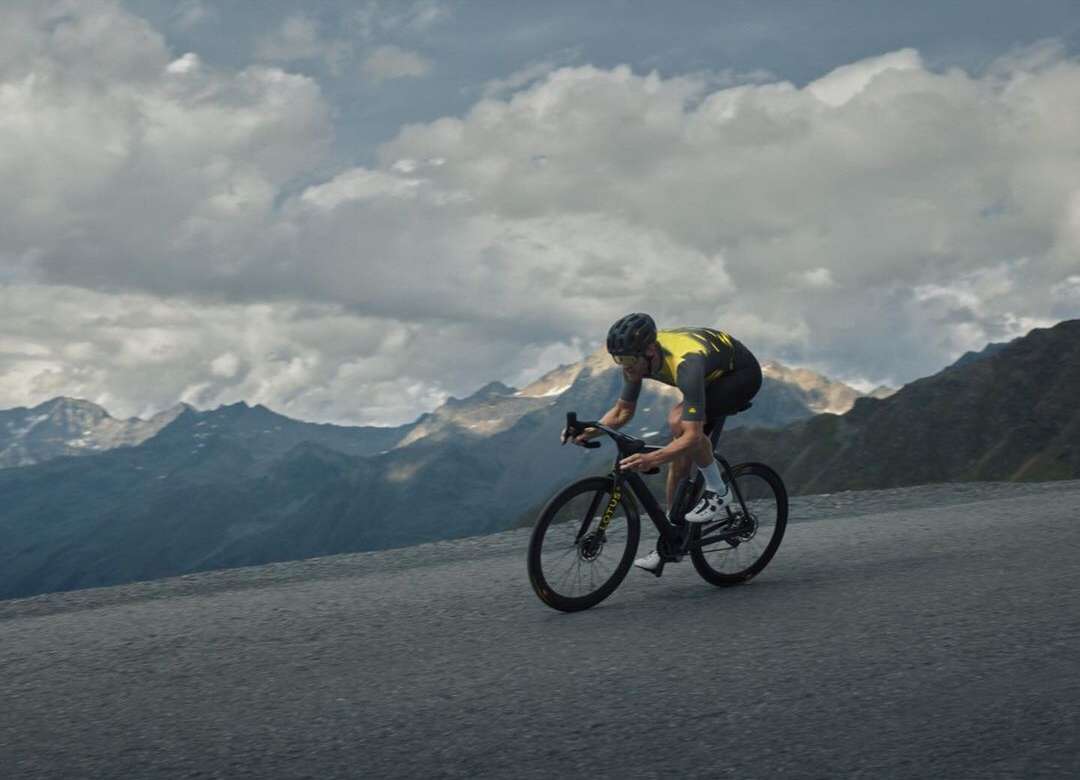 An adult male cyclist wearing helmet and sportswear rides a road bike uphill on asphalt, with mountain range and cloudy sky in background.