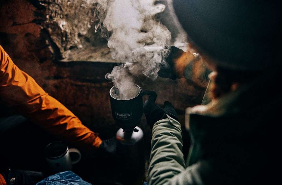 Camping scene with a person holding a steaming Primus mug in a dark, rocky environment, illuminated by warm light, creating a cozy ambiance.
