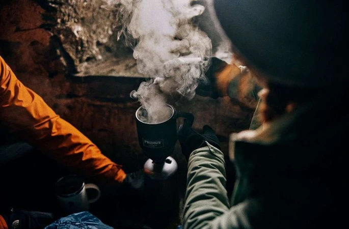 Camping scene with a person holding a steaming Primus mug in a dark, rocky environment, illuminated by warm light, creating a cozy ambiance.