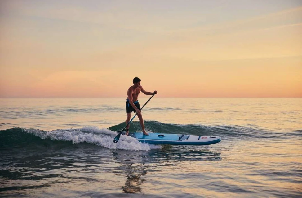 Man paddleboarding on ocean at sunset. Athletic man paddle surfing on blue board in tranquil sea environment with colorful sky background.