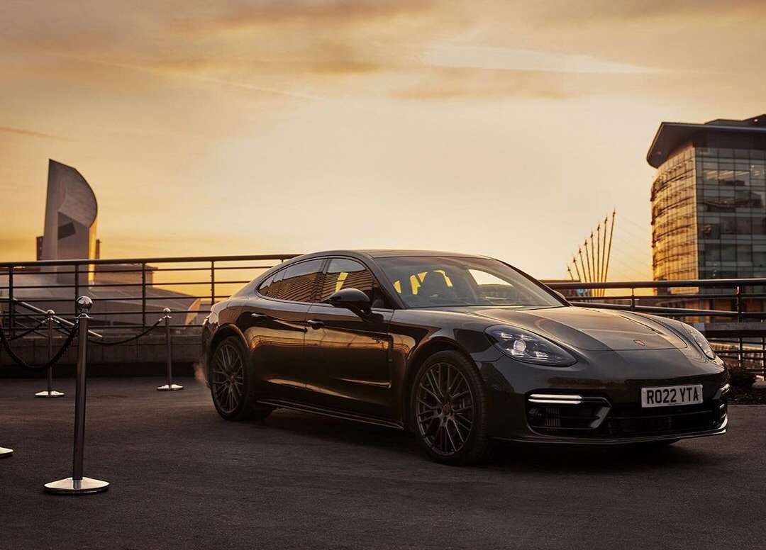 Sleek black Porsche Panamera parked on an outdoor surface with cityscape, architecture, and sunset sky providing a scenic backdrop.