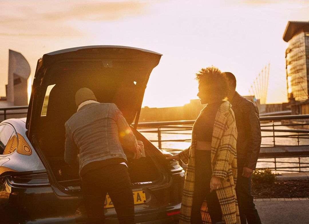Three adults stand by the open trunk of a car near a river at sunset with modern buildings in the background and golden light.