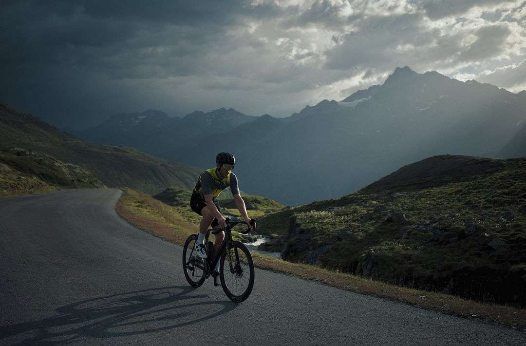 An adult male cyclist riding a road bike on a paved mountain road with overcast sky and mountain range backdrop in a fitness scene.