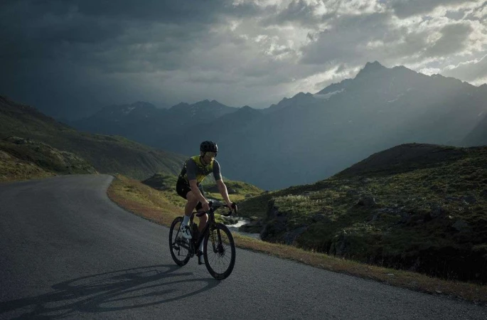 An adult male cyclist riding a road bike on a paved mountain road with overcast sky and mountain range backdrop in a fitness scene.
